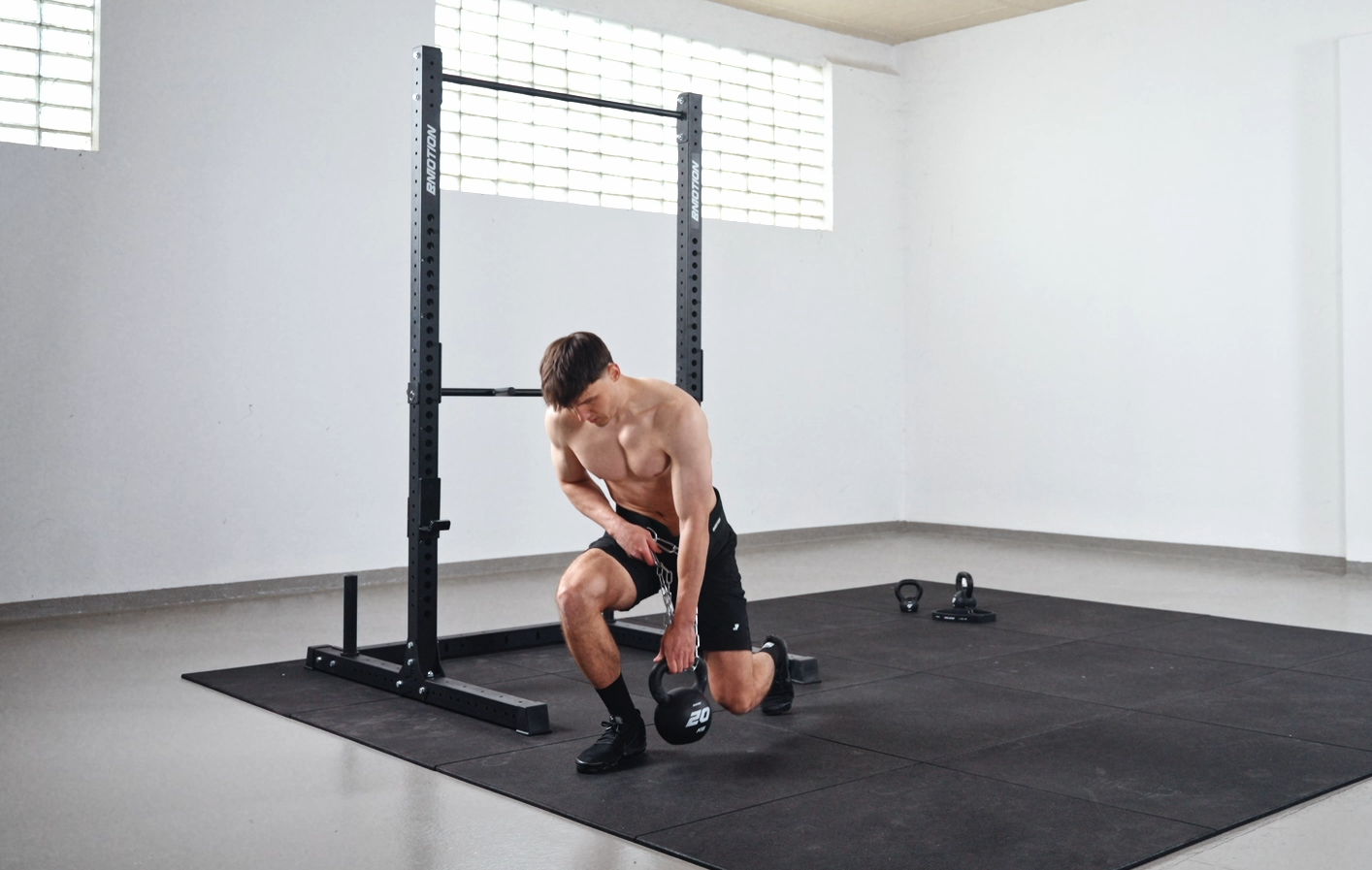 Athlet kniet im Fitnessstudio und befestigt eine Kettlebell an der Kette des Bmotion Gewichtsgürtels, im Hintergrund Power Rack Station